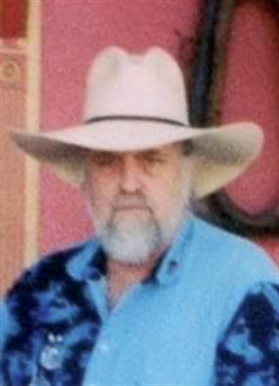A man wearing a cowboy hat stands outdoors, dressed in a blue shirt, with a vibrant background.