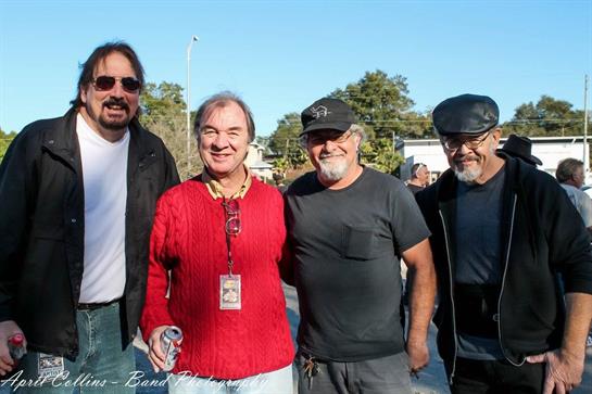Four musicians enjoy a sunny afternoon outdoors, socializing and smiling in a relaxed atmosphere.