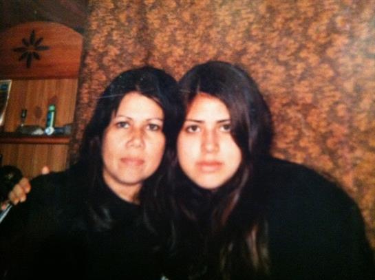 Two women with long dark hair sit closely together, smiling softly in a cozy indoor space.