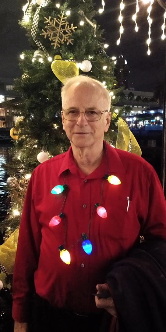 An elderly man in a red shirt stands by a brightly decorated tree by the water, smiling joyfully.