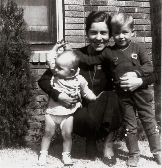A woman smiles while holding a toddler and standing beside a young boy in a garden.