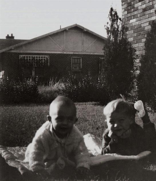 Two children enjoy playtime on a blanket outside a house surrounded by grass and plants.