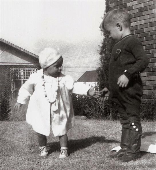 Two toddlers are exploring and holding hands in a sunny backyard filled with grass.