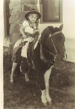 Child dressed in cowboy attire joyfully rides a small pony at a lively outdoor fair during the day.
