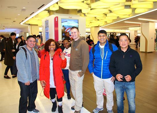 Friends gather and pose for a cheerful photo inside a lively shopping mall filled with people.
