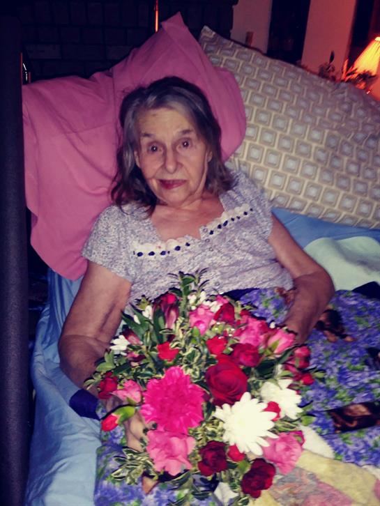 An elderly woman is resting in bed, holding a vibrant bouquet of flowers with a content expression.