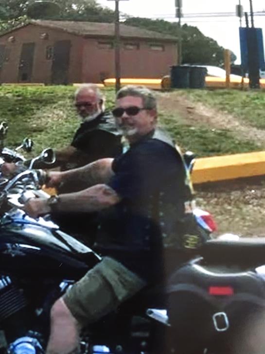 Two motorcyclists relax at a park, smiling and chatting while enjoying the sunny weather.