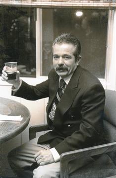 A man with a mustache enjoys a drink while seated at a table, dressed in a suit.