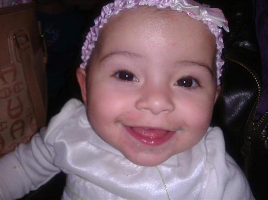 A joyful baby girl smiles brightly while wearing a white outfit and a cute headband indoors.