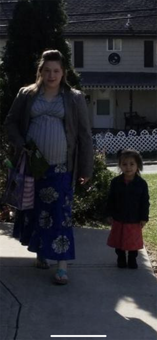 A pregnant woman stands outside with her young daughter, enjoying a sunny day in the neighborhood.