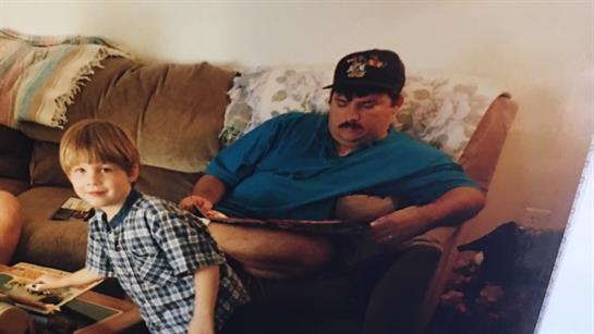 A boy plays nearby while his father relaxes on the couch with a magazine in hand.