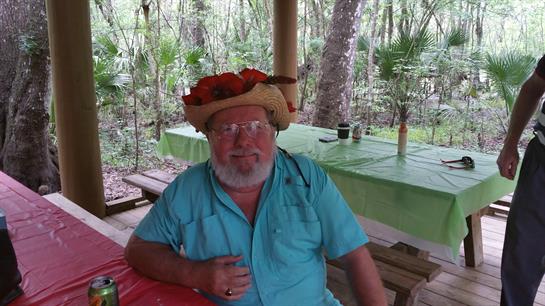 An elderly man smiles while seated at a table surrounded by greenery, wearing a colorful straw hat.