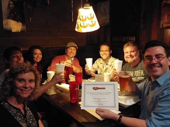 Friends gather around a table in a pub, raising glasses in celebration while holding a certificate.