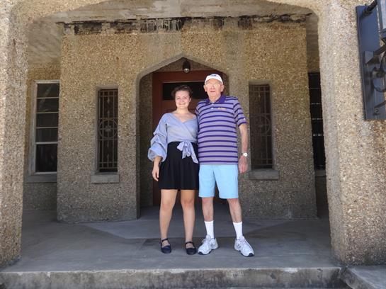 Two people stand in front of a textured stone structure, smiling joyfully under the sunlight.