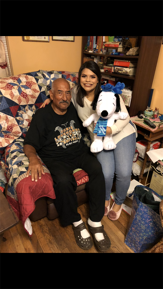 A woman and an elderly man smile together near cozy decor and a Snoopy toy.