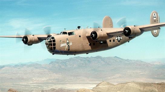A military plane flies gracefully above a mountainous desert terrain under bright sunlight.
