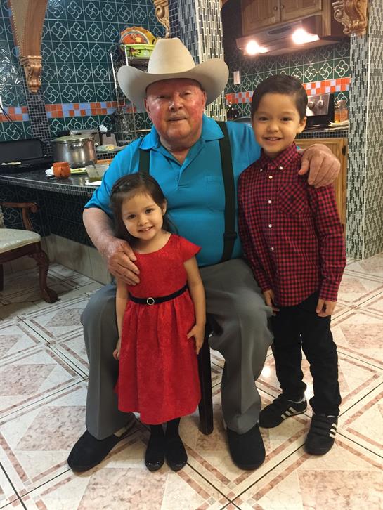 An elderly man enjoys quality time with his smiling grandchildren in a warm kitchen atmosphere.