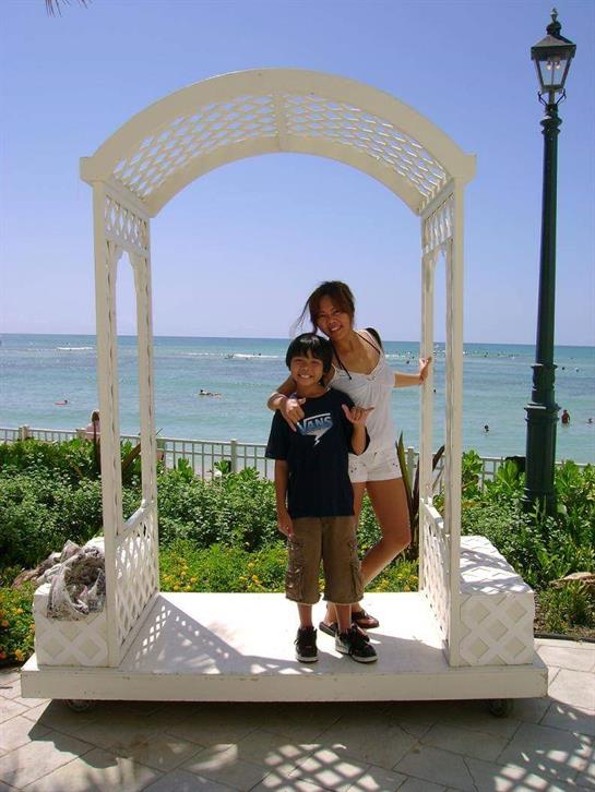 A young boy and a woman happily pose together by a white gazebo with a beach and ocean behind them.