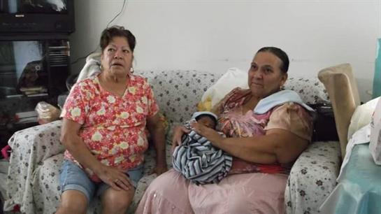 Two women sit on a couch, one cradling a newborn baby while the other looks on fondly.