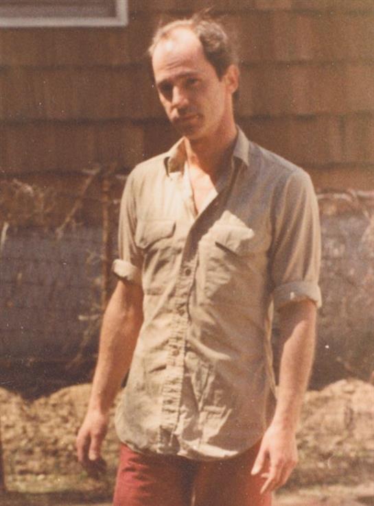 A young man poses outdoors in casual clothes amid a sunny rustic landscape.