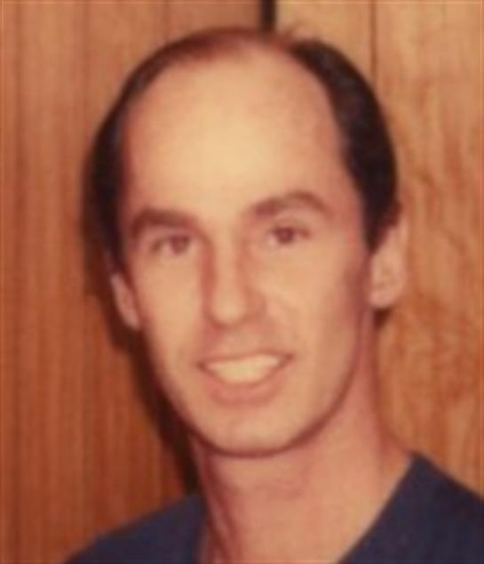 An individual smiles warmly while standing in front of wooden paneling, sharing a casual moment.