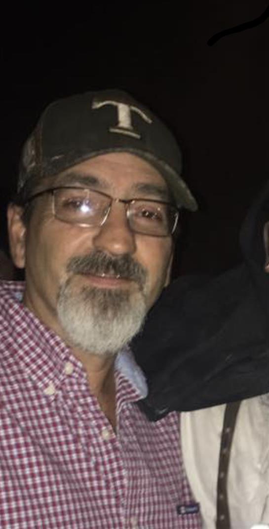 A man wearing a cap and glasses smiles warmly while socializing with friends in low light.