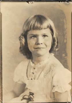 A young girl with a charming smile sits for a vintage-style portrait, wearing a light dress.