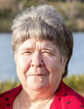 A senior woman stands smiling, basking in sunlight by a peaceful lake surrounded by greenery.