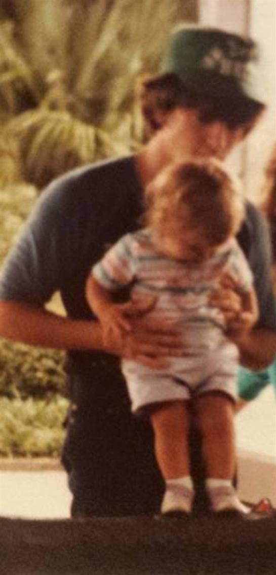 A parent lovingly holds a small child in a vibrant, sunlit backyard, conveying warmth and happiness.