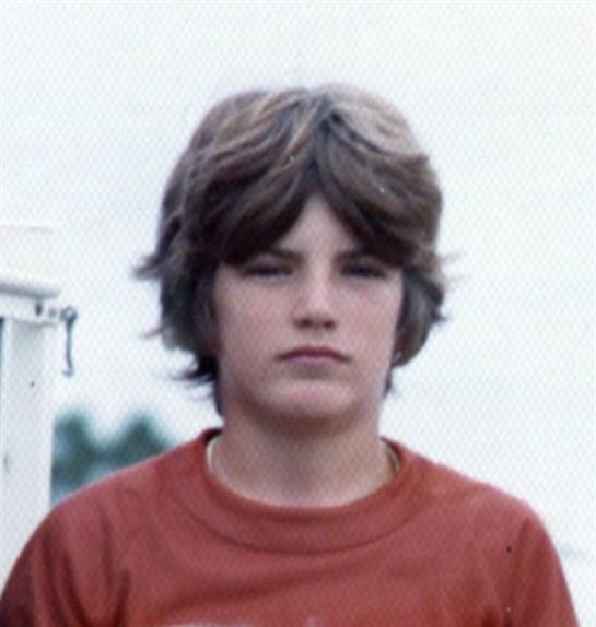 A young boy stands outdoors with somber expression in a red shirt, surrounded by nature.