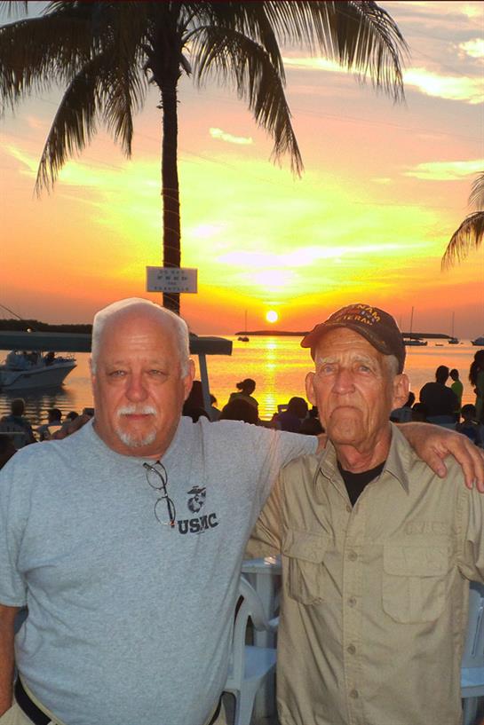Two men stand side by side against a vibrant sunset, surrounded by a lively waterfront scene.
