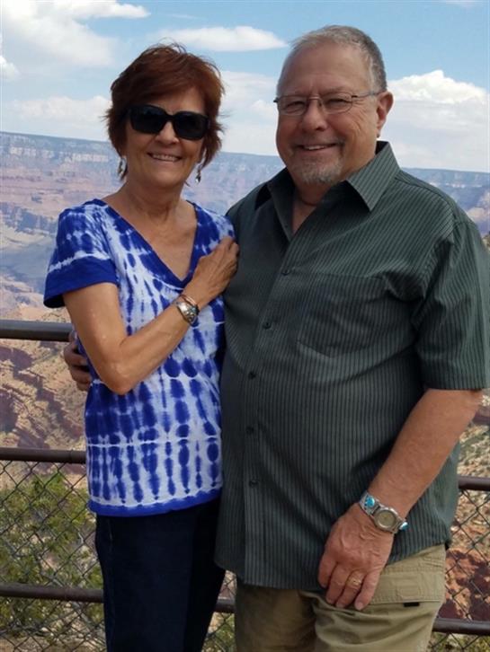 A couple smiles together while standing at the Grand Canyon overlook under a clear sky.