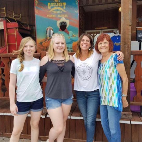 Four women and girls stand together, smiling in front of a colorful shop in a lively area.
