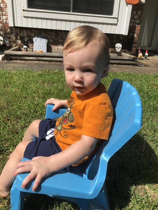 A young child sits on a blue chair in a backyard, playing amidst outdoor decorations on a sunny day.