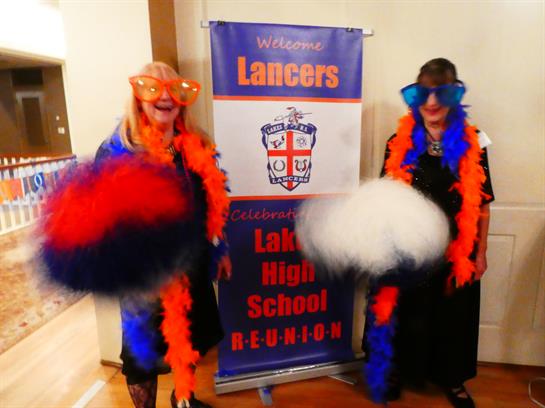Two alumni wear vibrant costumes and pose with pom-poms at a high school reunion event.