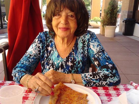 An older woman smiles while savoring a slice of pizza at a lively outdoor location.