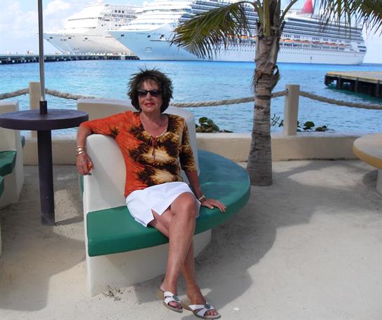 A woman relaxes by the water, sitting on a bench with a cruise ship behind her.