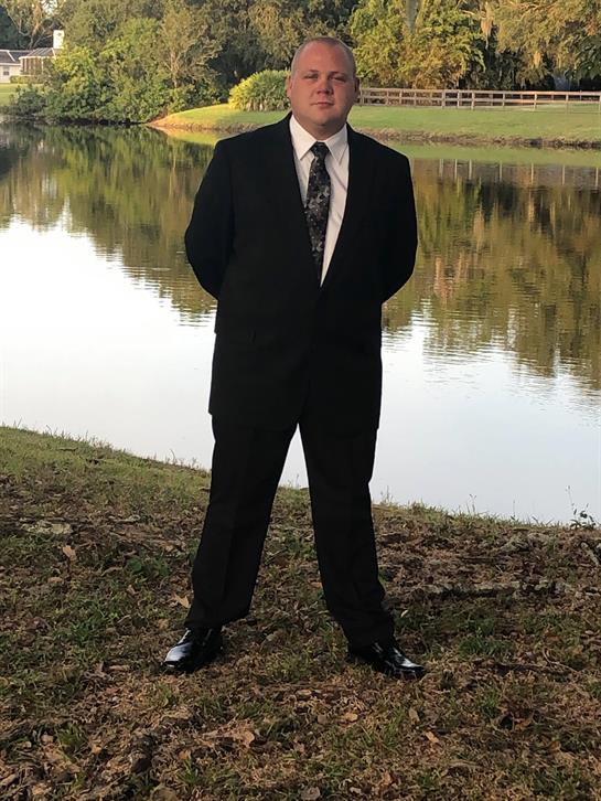Individual in formal suit stands confidently by a calm lake reflecting the natural scenery.