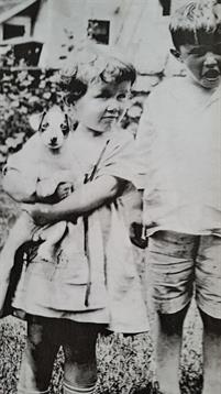 Two children stand together in a garden, one holding a small puppy in their arms.
