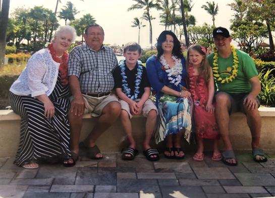 A family poses together wearing colorful leis, smiling under the warm Hawaiian sun near palm trees.