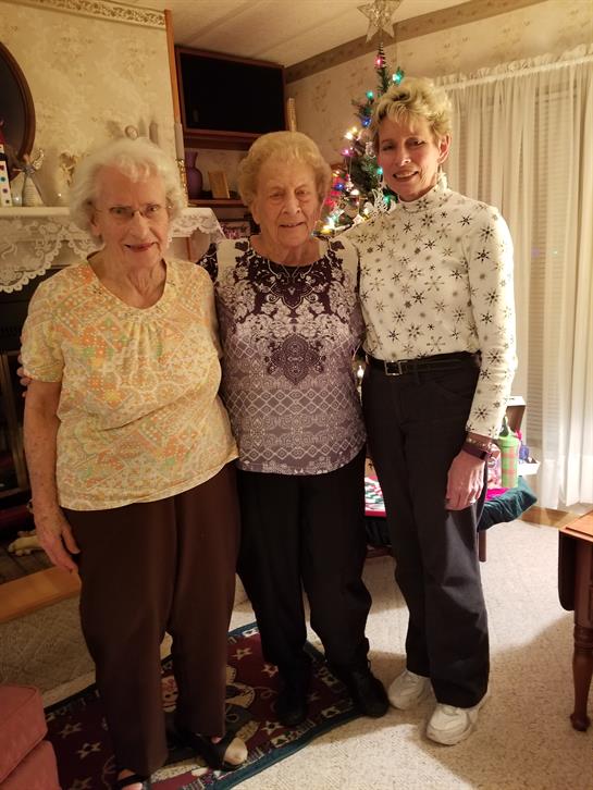 Three smiling elderly women stand together in a warm living room decorated for a holiday gathering.