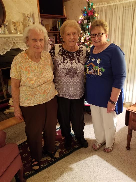 Three women stand together smiling in a comfortable living room decorated for a festive celebration.