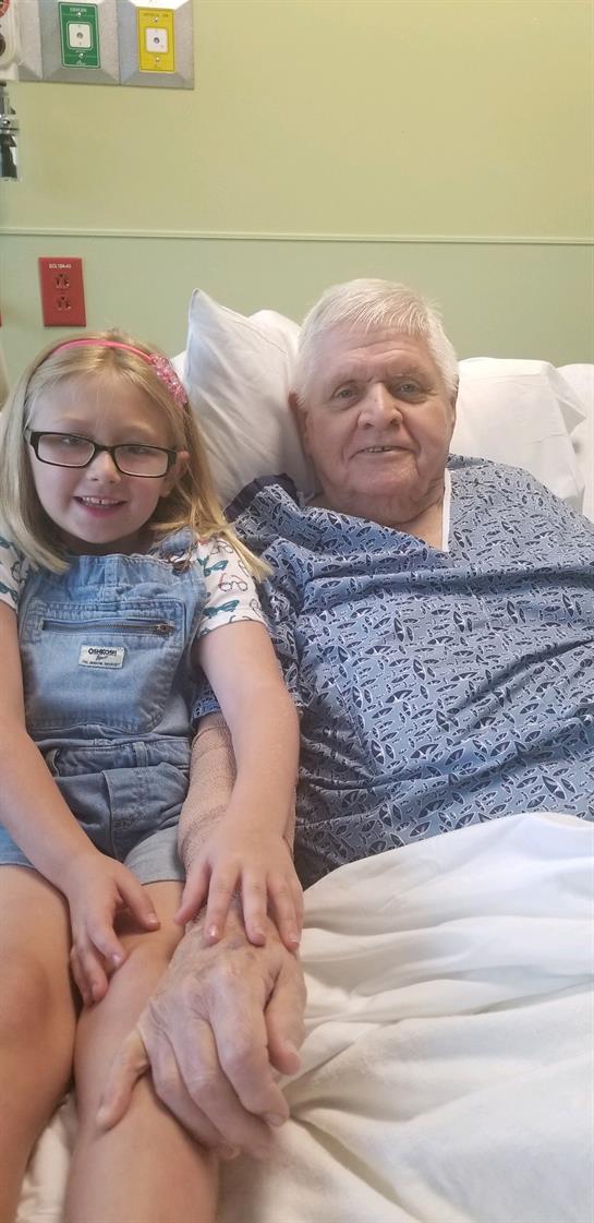 A young girl in glasses smiles while sitting beside her grandfather in a hospital bed.