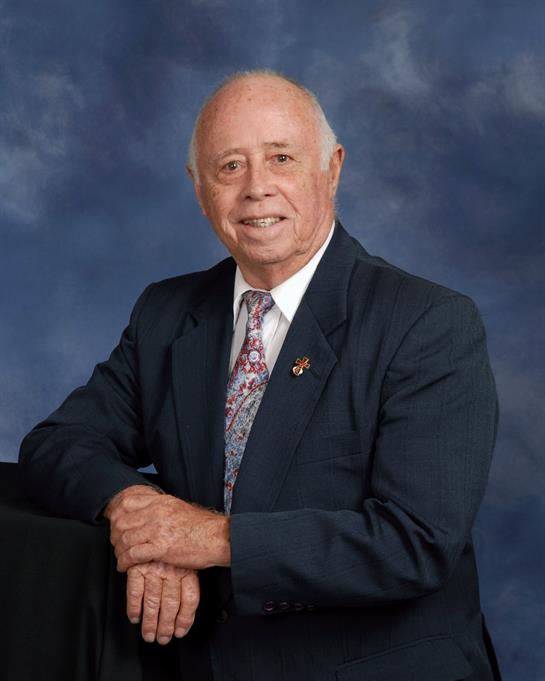 An elderly man with gray hair is smiling confidently while wearing a dark suit and tie in a studio.