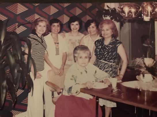 A group of six women pose closely together while one child sits at a table in a cheerful space.
