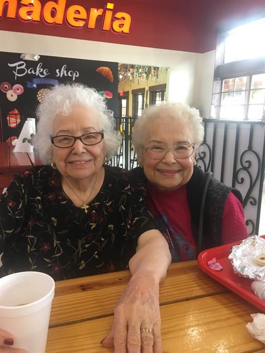 Two senior women sit together at a table in a bake shop, enjoying each other's company and treats.