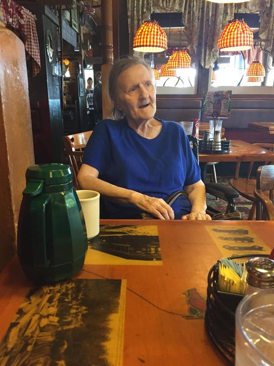An elderly woman in a blue shirt sits at a cafe table, enjoying a drink while conversing.