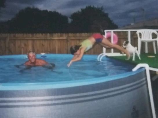A child joyfully leaps into a backyard pool while an adult watches, enjoying a summer evening.