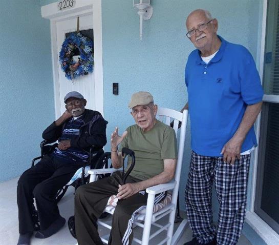 Three elderly men sit and stand on a porch, sharing laughs and enjoying their time together.