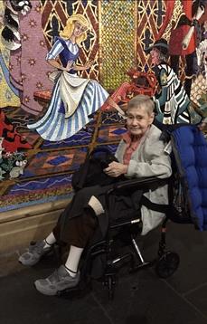 A woman sits in a wheelchair, smiling as she admires vibrant art displayed on the walls.